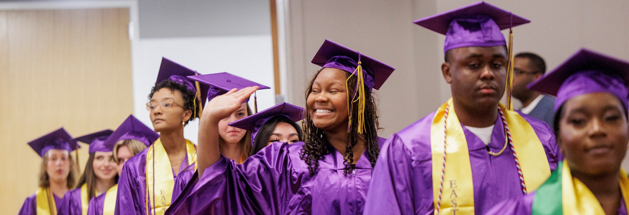 HHP graduates smile and wave while walking in purple and gold caps and gowns.