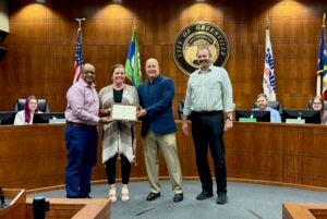 College professor holds an award certificated while standing with 3 others in a City Hall room.
