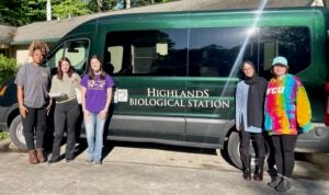 Five college students stand outside a green bus.