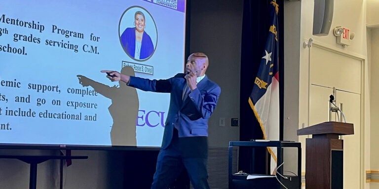 Two college faculty members stand and present. One of them, a male, is pointing at projector screen