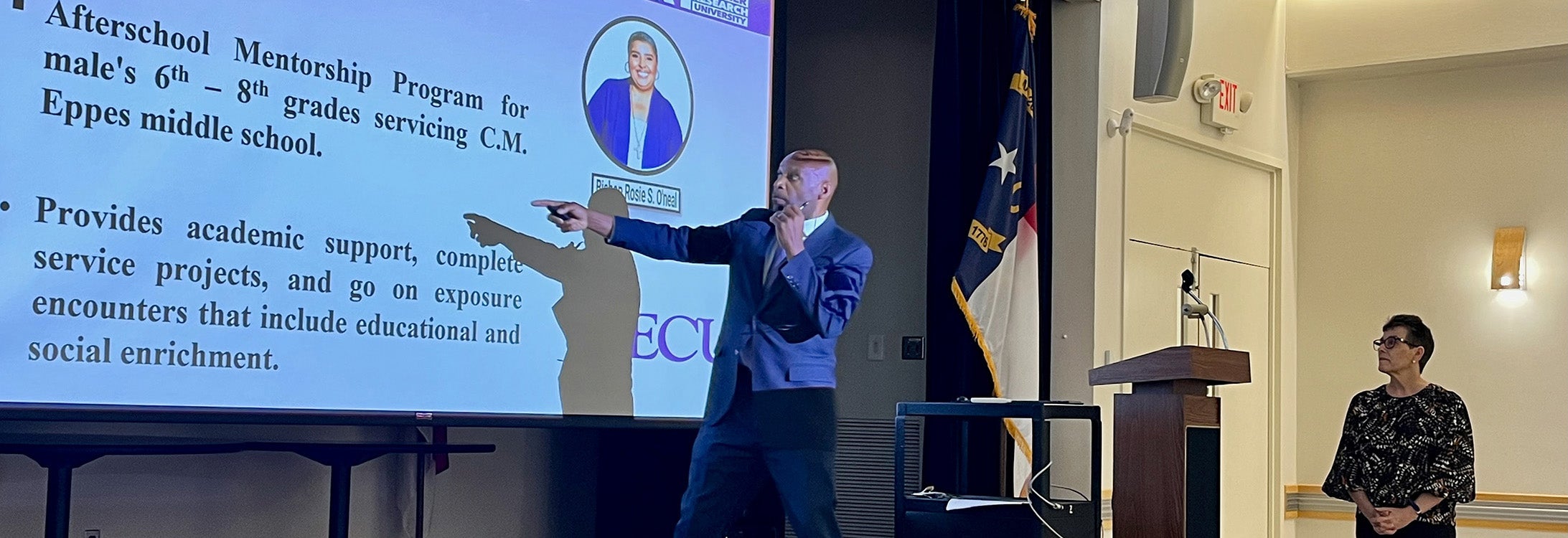Two college faculty members stand and present. One of them, a male, is pointing at projector screen