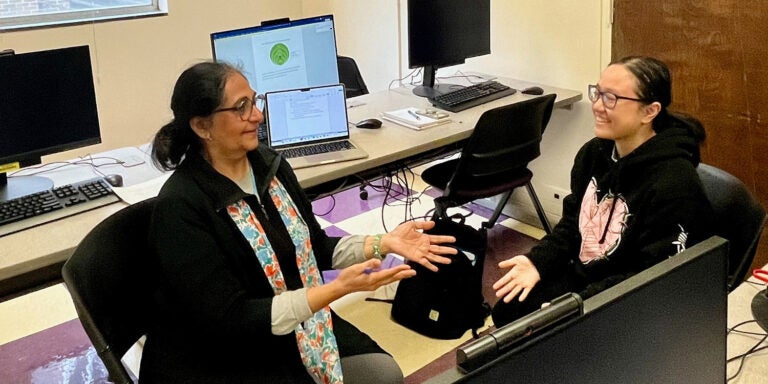 Professor and student sitting and talking in computer lab with computer screens in background.