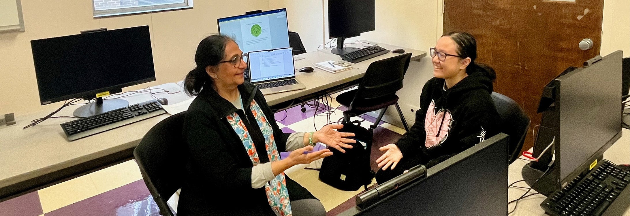 Professor and student sitting and talking in computer lab with computer screens in background.