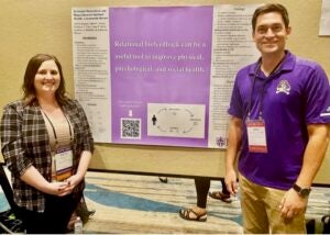 A student, Adrian Knauss, stands with faculty member Jake Jensen at a purple and a white poster.
