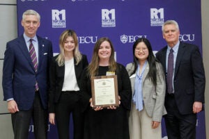 A college professor holds an award plaque while standing in a group in front of a purple research banner.