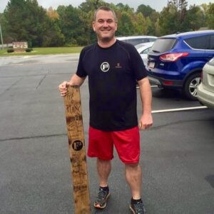A man stands in a parking lot while holding a wooden plank beside him.