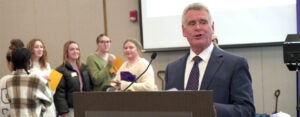 A man wearing a suit and tie stands at a podium andspeaks while six young students stand in the background.