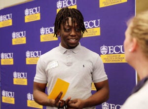 A young man wearing a gray polo shirt smiles as hespeaks to a woman standing at right.